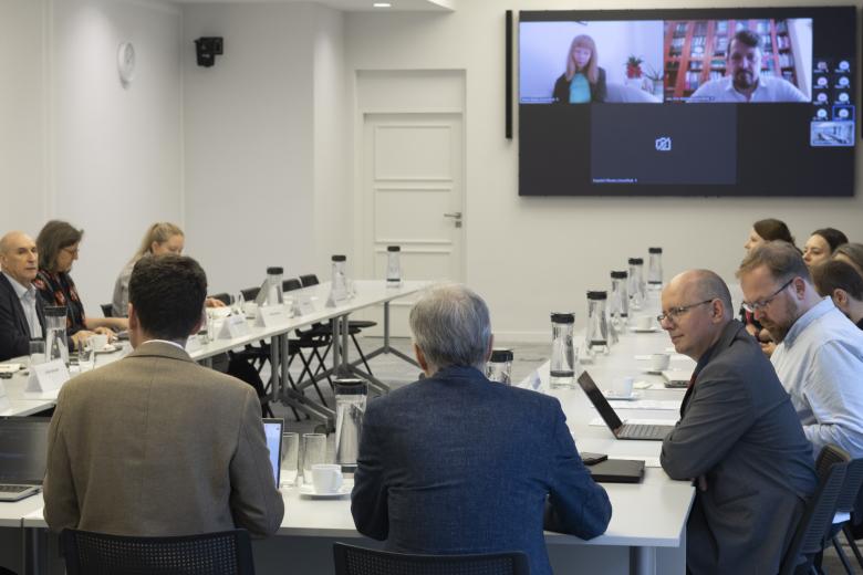 Kilkanaście osób rozmawia siedząc przy stołach konferencyjnych ustawionych w kształt podkowy. W szczycie stołu znajduje się ekran wyświetlający osoby uczestniczące w spotkaniu za pośrednictwem internetu.