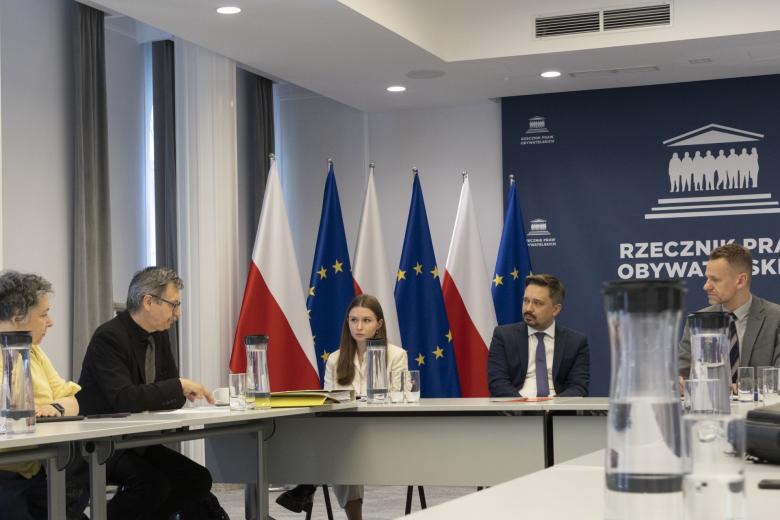 Osoby siedzące za stołem w sali konferencyjnej, w tle flagi Polski i Unii Europejskiej