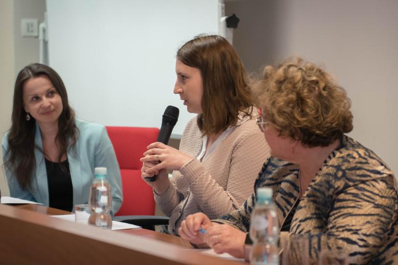 Trzy osoby siedzą przy stole, jedna z nich trzyma w ręku mikrofon i wypowiada się