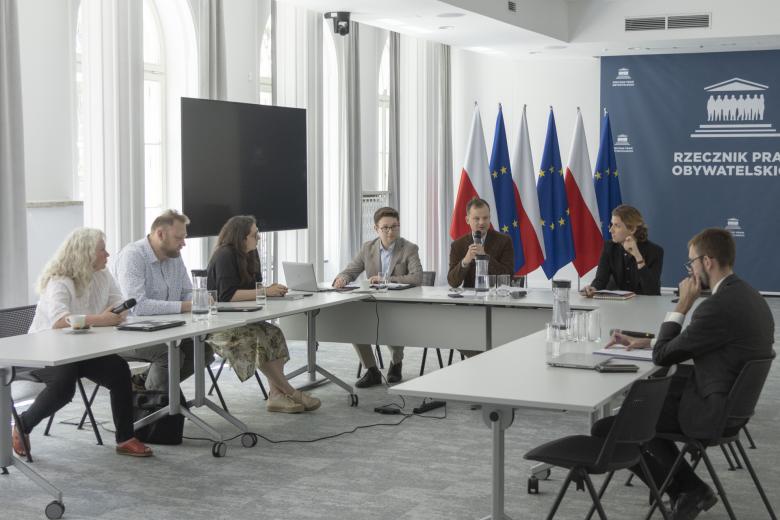Kilka osób siedzi za stołami ustawionymi w podkowę w sali konferencyjnej