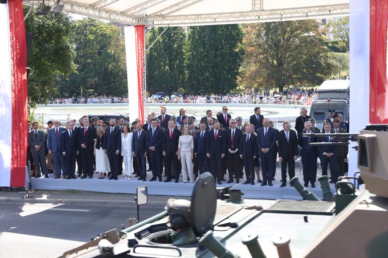 Kilkadziesiąt osób stoi na zadaszonej i przyozdobionej polskimi flagami trybunie. Przed trybuną przejeżdża opancerzony pojazd wojskowy. Za trybuną widać tłum ludzi