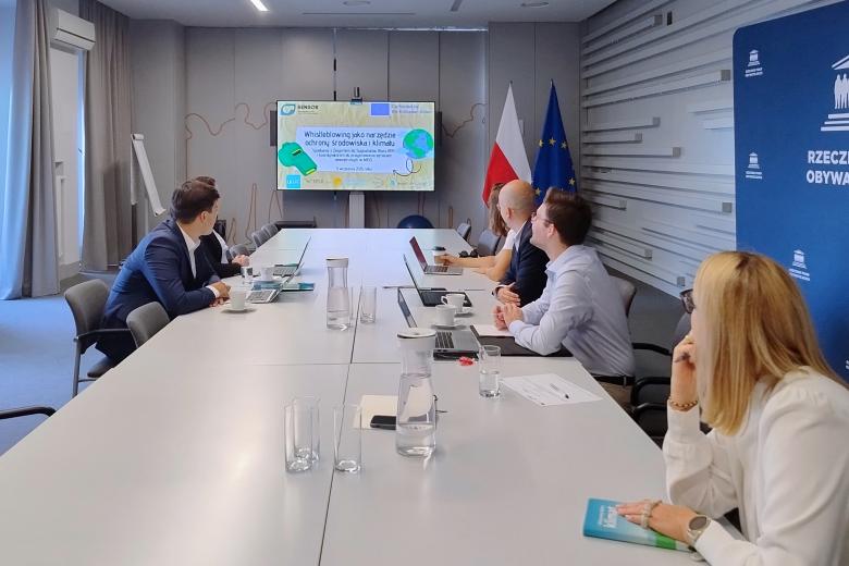 Sześć osób siedzi przy stole konferencyjnym i patrzy na prezentację wyświetlaną na ekranie stojącym w szczycie stołu