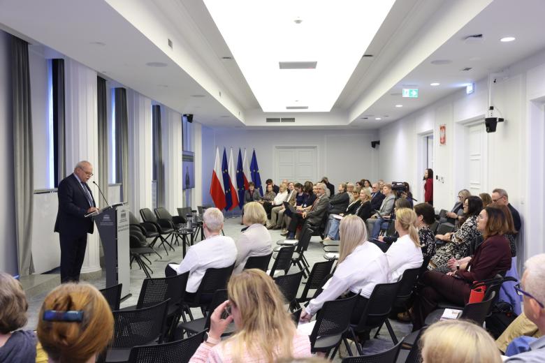 Kilkadziesiąt osób na sali konferencyjnej siedzi i słucha wystąpienia osoby mówiącej za mównicą