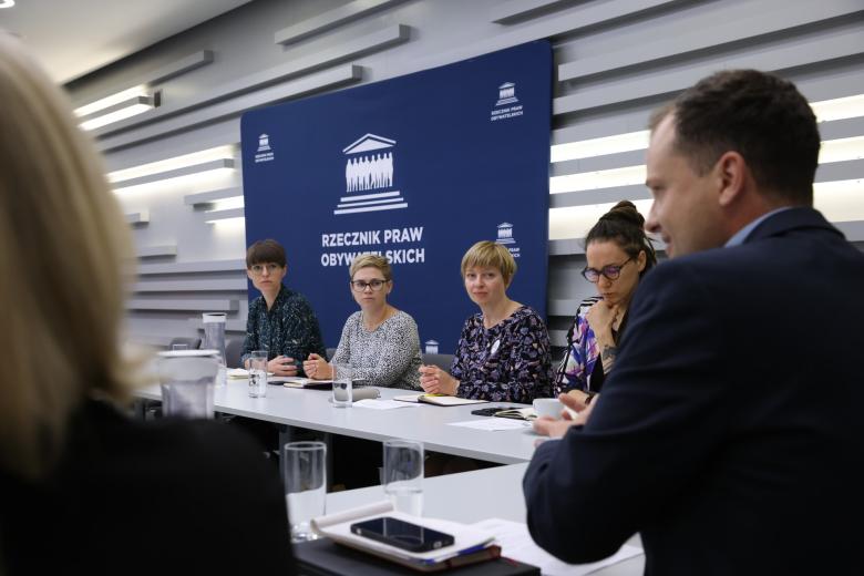 Kilka osób siedzi za stołem w sali konferencyjnej, za nimi duża ścianka z białym napisem "Rzecznik Praw Obywatelskich" na granatowym tle