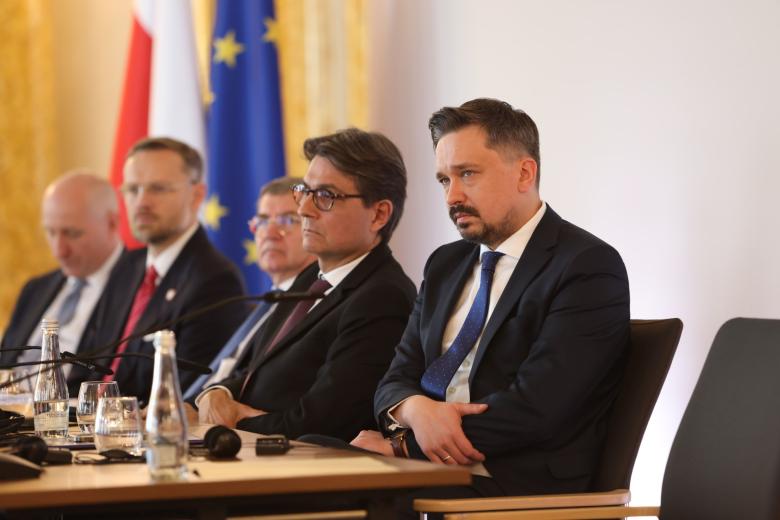 Kilka osób siedzi za podłużnym stołem w bogato zdobionej, zabytkowej sali