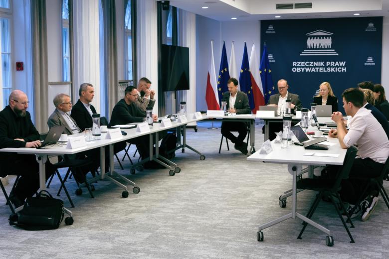 Kilkanaście osób siedzi przy stołach ustawionych w podkowę, za stołami przy ścianie rząd flag Polski i UE