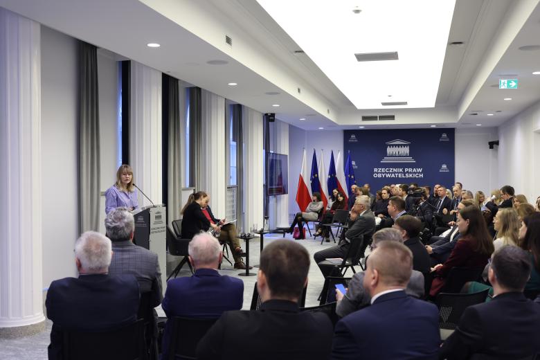 Dominika Czerniak wypowiada się stojąc przy mównicy w sali konferencyjnej. Wokół niej na widowni siedzi kilkadziesiąt osób