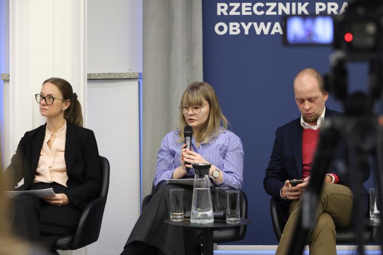 Trzy osoby uczestniczące w panelu siedzą obok siebie na krzesłach