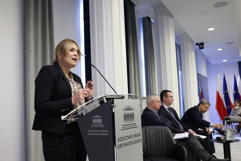 Barbara Janusz-Pohl wypowiada się stojąc przy mównicy w sali konferencyjnej. Obok siedzą trzy osoby - uczestnicy panelu