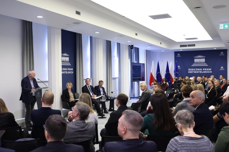 Krzysztof Bondaryk wypowiada się stojąc przy mównicy w sali konferencyjnej. Dookoła niego na widowni siedzi kilkadziesiąt osób