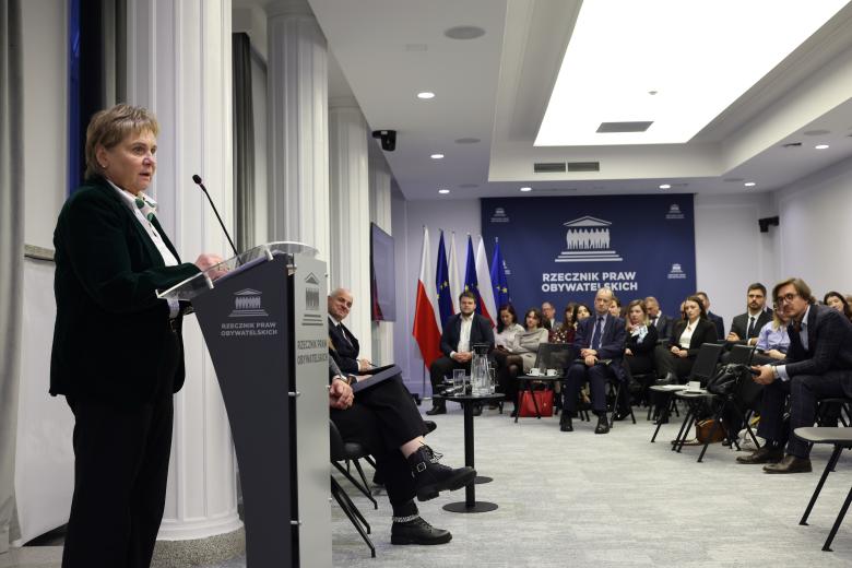 Beata Najjar wypowiada się stojąc przy mównicy w sali konferencyjnej. Wokół na widowni siedzi kilkadziesiąt osób