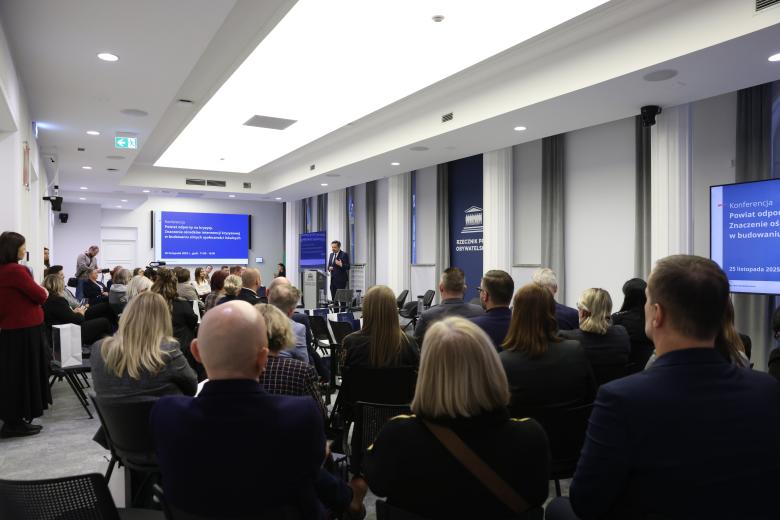 Marcin Wiącek wypowiada się stojąc przy mównicy. Wokół niego na widowni siedzi kilkadziesiąt osób