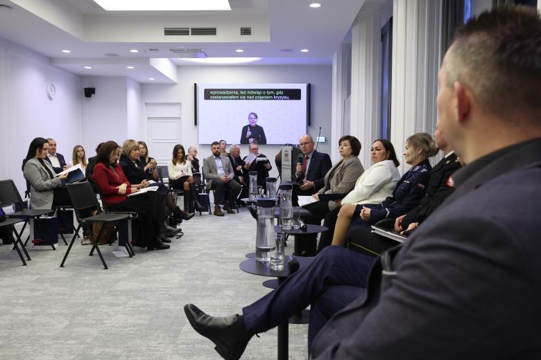 Kilka osób rozmawia siedząc na krzesłach ustawionych w szeregu. Naprzeciwko nich na widowni siedzą inni uczestnicy konferencji