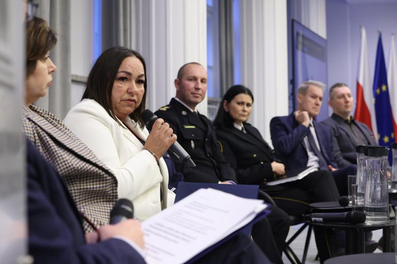 Uczestnicy panelu siedzą na krzesłach ustawionych w szeregu. Jedna z uczestniczek wypowiada się trzymając mikrofon