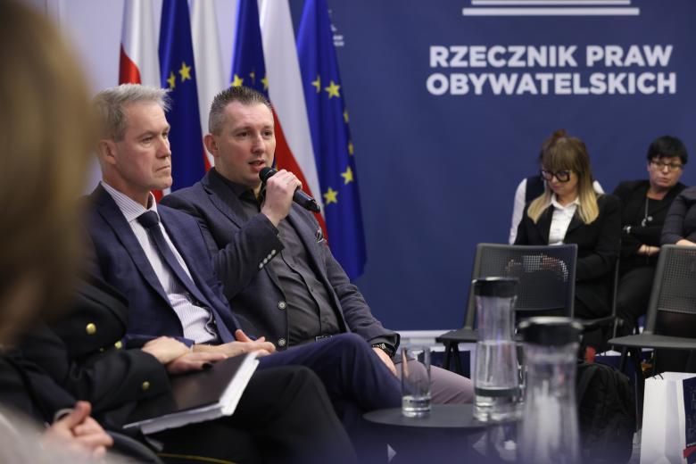 Dwóch uczestników panelu siedzi obok siebie. Jeden z nich trzyma mikrofon i się wypowiada. Naprzeciwko nich na widowni siedzą inni uczestnicy konferencji