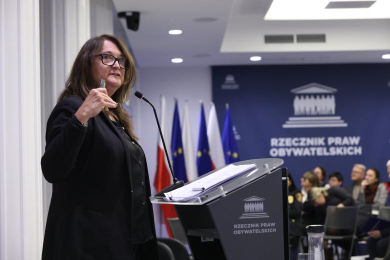 Beata Karlińska wypowiada się stojąc przy mównicy. W tle na widowni widać sylwetki siedzących innych uczestników konferencji