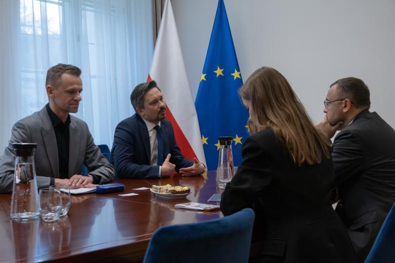 Cztery osoby siedzą przy stole i rozmawiają w salce konferencyjnej. Obok stołu stoją flagi Polski i Unii Europejskiej