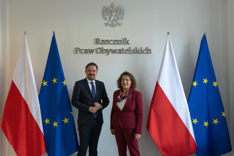 RPO Marcin Wiącek i Melinda Simmons uśmiechają się pozując do zdjęcia na tle flag Polski i Unii Europejskiej oraz godła Polski i napisu "Rzecznik Praw Obywatelskich"
