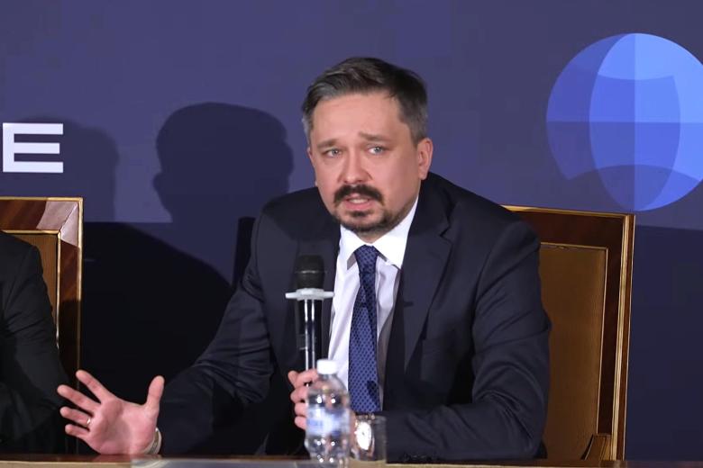 Osoba siedząca za stołem mówi do trzymanego w dłoni mikrofonutrzymając w dłoni mikrofo