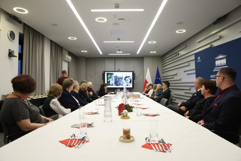 Kilkanaście osób siedzi przy dużym stole w sali konferencyjnej, słucha wystąpienia i przygląda się prezentacji wyświetlanej na dużym ekranie