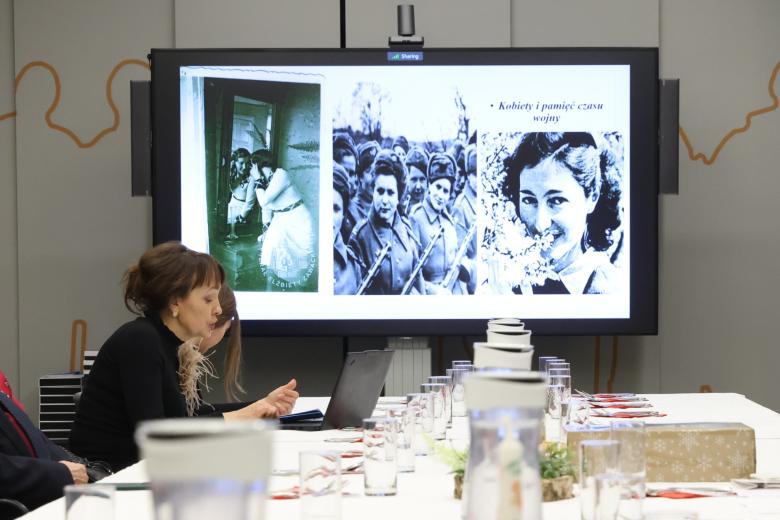 Kobieta cos opowiada na tle wyświetlacza z widoczną prezentacją. Na prezentacji czarno-białe zdjęcia kobiet i napis "Kobiety i pamięć czasu wojny"
