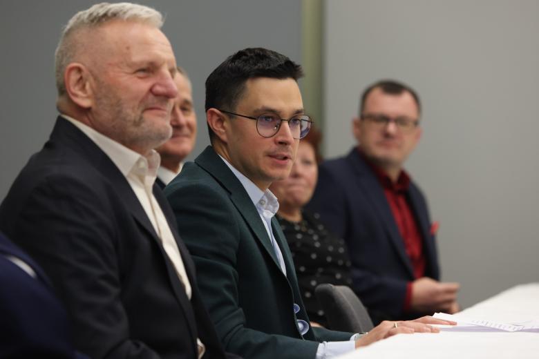 Kilka osób siedzi przy stole, na pierwszym planie dwóch mężczyzn w garniturach, jeden z nich coś mówi