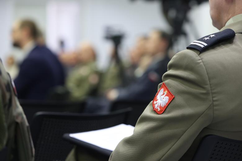 Siedząca sylwetka człowieka w mundurze. Na ramieniu munduru jest naszywka z godłem Polski - orłem w koronie