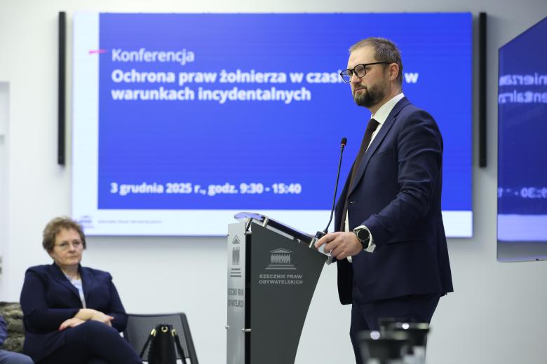 Tomasz Oklejak stoi przy mównicy w sali konferencyjnej. W tle widać wyświetlacz, na którym jest nazwa konferencji