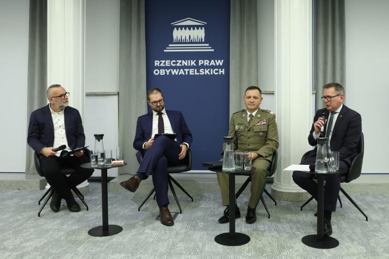 Cztery osoby rozmawiają siedząc obok siebie na krzesłach w sali konferencyjnej.