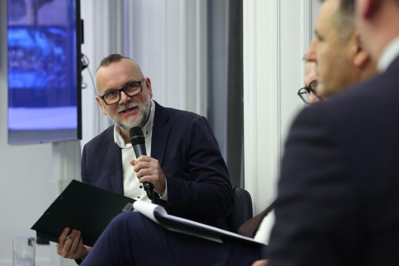 Męzczyzna siedzi na krześle i wypowiada się w stronę siedzących przed nim innych uczestników panelu 