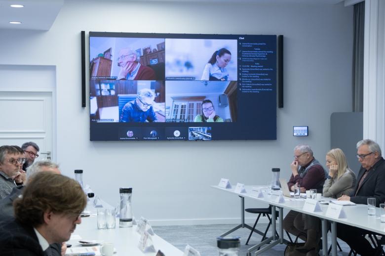 osoby siedzą w sali w tle duży ekran ze zdjęciami osób