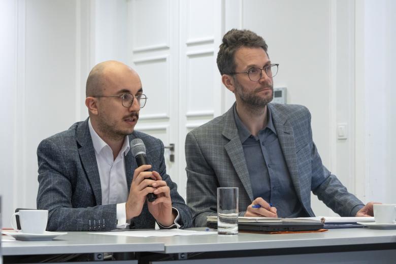 dwie osoby siedzą za  stołem jedna mówi do mikrofonu