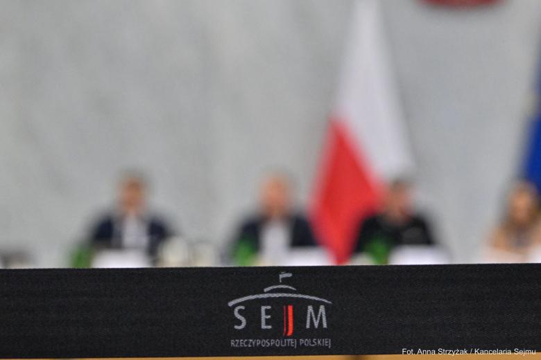 osoby siedzą w sali z polską flagą 