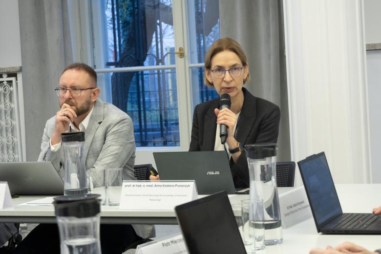 dwie osoby siedzą za stołem w sali jedna mówi do mikrofonu  