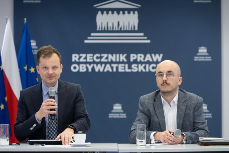 dwie osoby siedzą za stołem jedna trzyma w dłoni mikrofon
