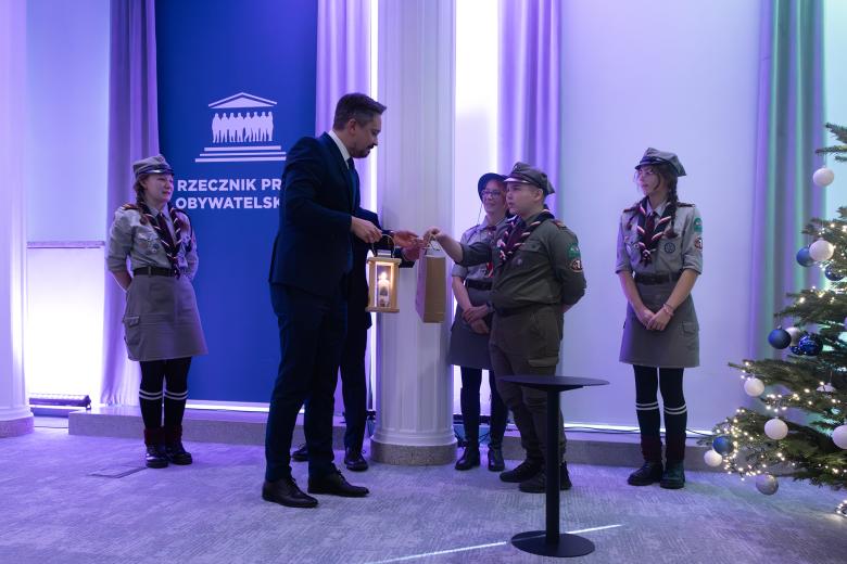 Marcin Wiącek przyjmuje od młodego harcerza lampion z zapaloną świecą. Przygląda się temu trzech innych harcerzy. Obok stoi udekorowana choinka