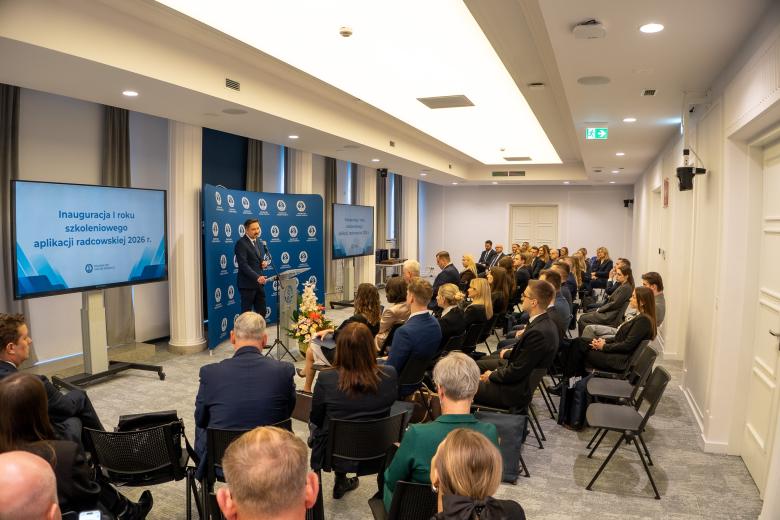 Kilkadziesiąt osób siedzi na sali konferencyjnej i słucha wystąpienia osoby mówiącej za mównicą na środku