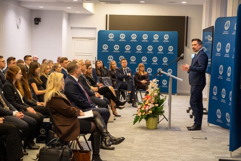 Kilkadziesiąt osób siedzących na sali konferencyjnej słucha wystąpienia osoby mówiącej do mikrofonu za mównicą