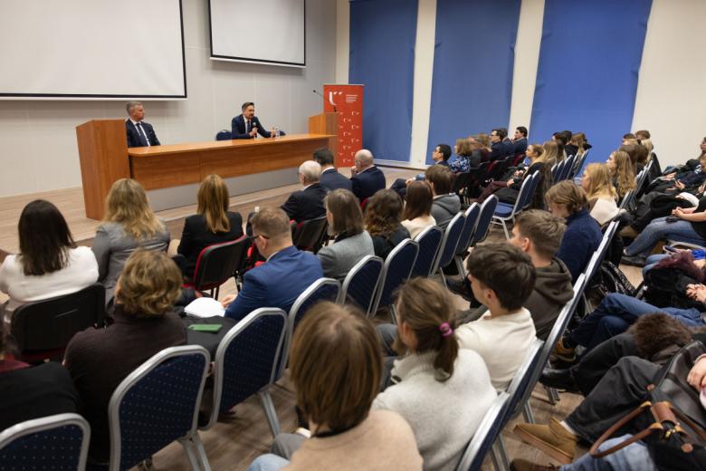 Osoba przemawia siedząc za stołem na sali wykładowej, przysłuchują jej się ludzie siedzący na krzesłach