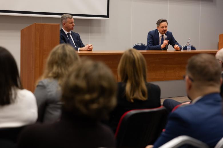 Osoba siedzi za stołem na sali wykładowej i mówi do trzymanego w ręku mikrofonu