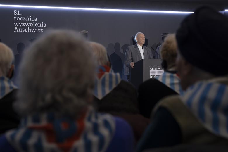 widziane od tyłu osoby siedzą w sali jedna w tle przemawia