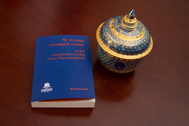 Na stole stoi bogato zdobiona, pozłacana misa. Obok niej leży książka o tytule "W stronę równych szans. 35 lat urzędu Rzecznika Praw Obywatelskich"