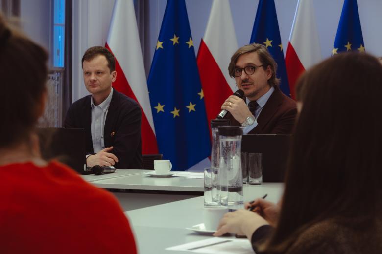 dwie osoby siedzą w sali jedna  przemawia 