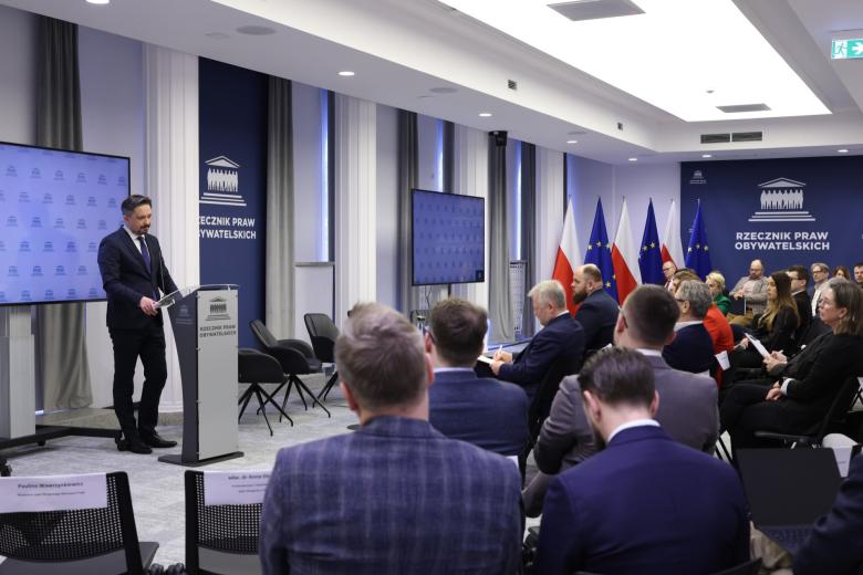 Kilkadziesiąt osób siedzi na sali konferencyjnej i słucha wystąpienia osoby mówiącej za mównicą na środku