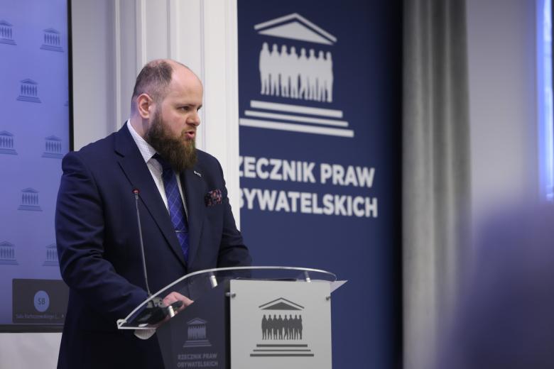 Osoba przemawia za mównicą na tle napisu "Rzecznik Praw Obywatelskich"