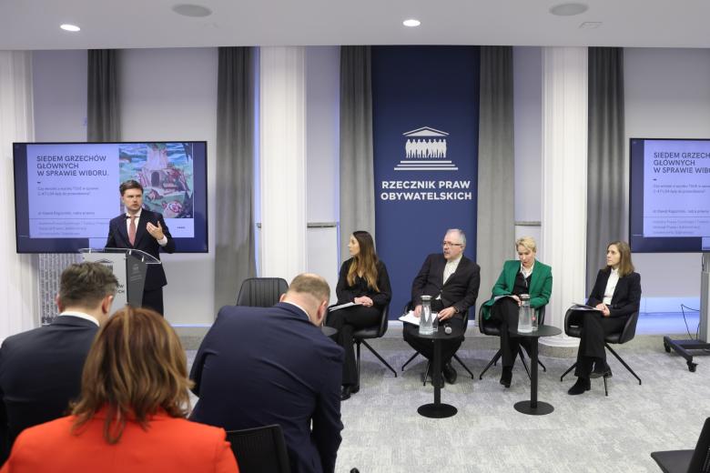 Cztery osoby siedzą na krzesłach, a jedna przemawia za mównicą w sali konferencyjnej