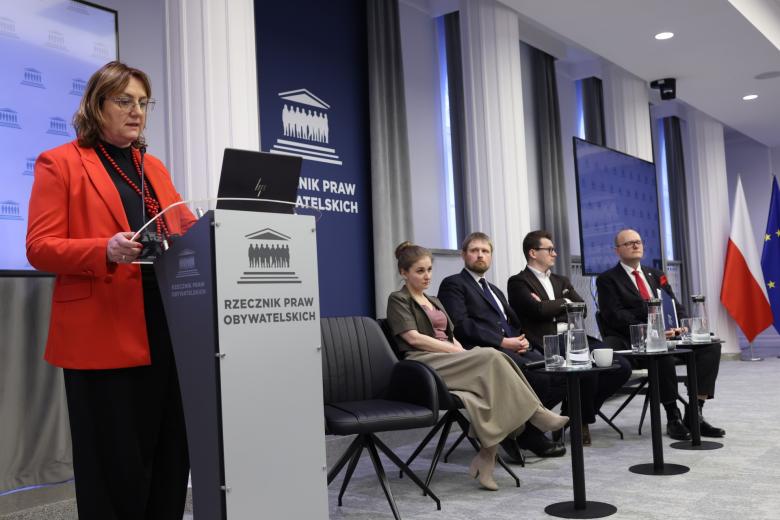 Osoba przemawia za mównicą, a obok cztery osoby siedzą na krzesłach i słuchają