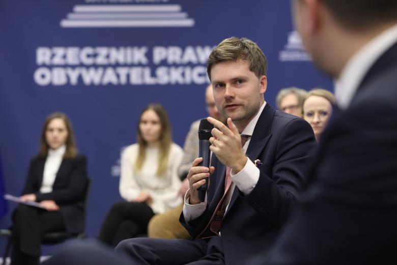 Osoba siedząca na krześle na sali konferencyjnej wypełnionej ludźmi mówi do mikrofonu trzymanego w dłoni