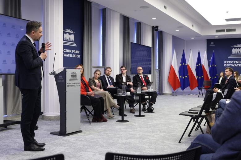 Osoba stoi i mówi do mikrofonu gestykulując na sali konferencyjnej wypełnionej słuchającymi ludźmi