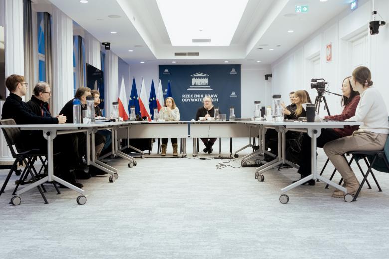 Kilkanaście osób siedzi przy stołach ustawionych w podkowę na sali konferencyjnej